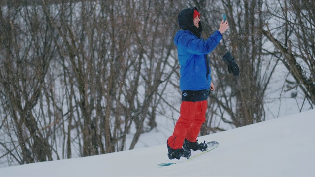 一名男性单板滑雪运动员骑着滑板到雪坡上，并在智能手机上给你的朋友发信息视频素材