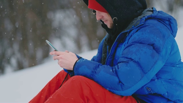 男滑雪板坐在雪地上用手机拍摄度假村的美丽风景，用于社交网络视频素材