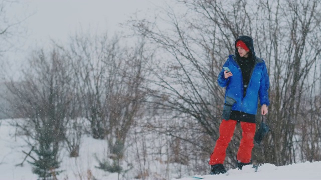 一名男性单板滑雪运动员骑着滑板到雪坡上，并在智能手机上给你的朋友发信息视频素材