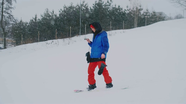 一名男性单板滑雪运动员骑着滑板到雪坡上，并在智能手机上给你的朋友发信息视频素材