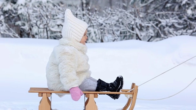 穿着白色毛皮大衣的小女孩坐在雪橇上，她在冬天的树林里打滚视频素材