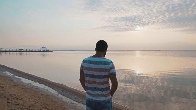 一个年轻人站在死海的岸边，欣赏美丽的日出。美丽的海景黎明。视频素材