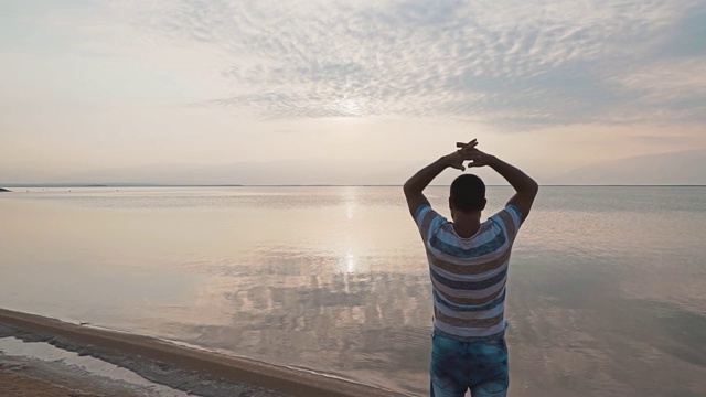 一个年轻人站在死海的岸边，欣赏美丽的日出。美丽的海景黎明。视频素材