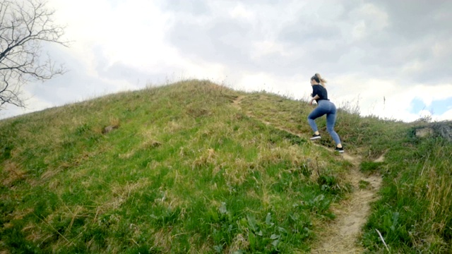 一个年轻女子跑上长满青草的小山。慢动作跑步，慢跑慢动作，美丽的云朵，运动女性户外运动视频素材