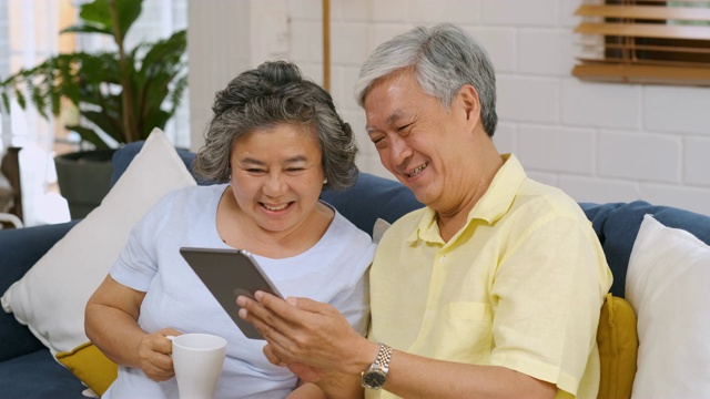 在家里的客厅里，一对亚洲老年夫妇坐在沙发上用桌子计算和喝咖啡。拥有科技生活方式的老年人。居家养老的概念。俯视图视频素材