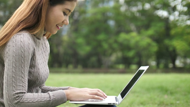 一个年轻的亚洲女人在公园里用笔记本电脑打字视频素材