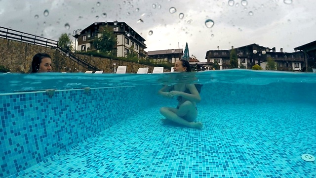 快乐的女孩们在雨天享受游泳池的雨滴击中水面，gopro圆顶半水下的景色，慢动作视频素材