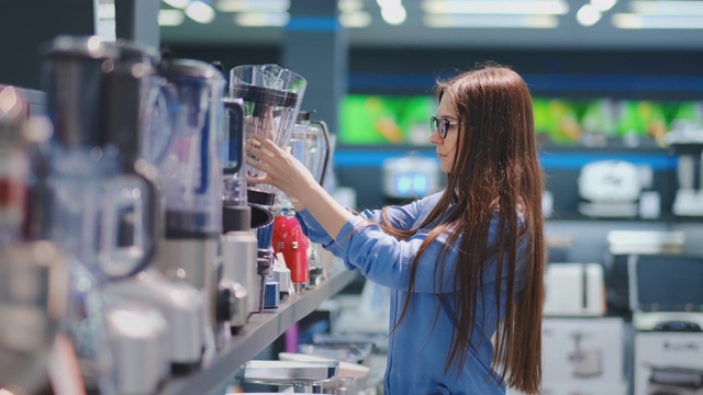 在家电商店里，一位身穿衬衫的黑发女子通过观看并拿在手里挑选搅拌机来购物视频素材