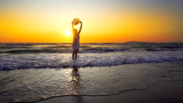 微笑自由和幸福的女人在海滩上日落。她在旅游、度假、户外享受着宁静的海洋大自然。白色礼服和帽子视频素材