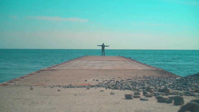 在大海和天空的背景下，一个年轻人站在码头上，双臂向天空举起。视频素材