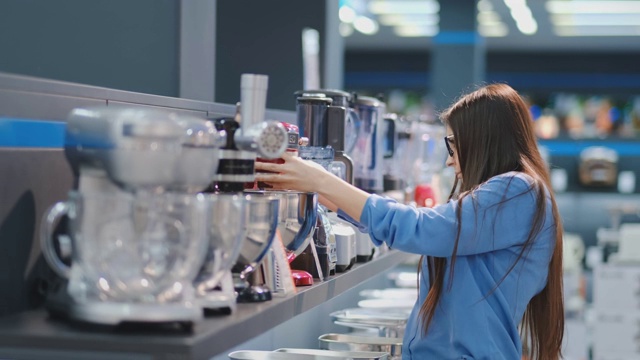 在家电商店里，一位身穿衬衫的黑发女子通过观看并拿在手里挑选搅拌机来购物视频素材