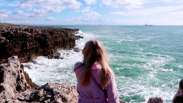 一个年轻女子坐在悬崖上看着汹涌的海浪。在晴朗的天气里，强烈的海浪撞击着岩石。纯粹的力量，自然力量的展示。海洋能源视频素材