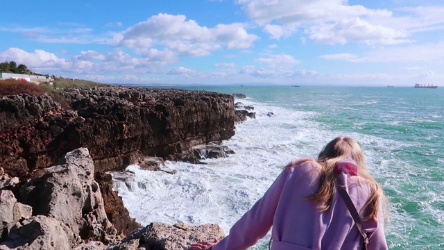 一个年轻女子坐在悬崖上看着汹涌的海浪。在晴朗的天气里，强烈的海浪撞击着岩石。纯粹的力量，自然力量的展示。海洋能源视频素材