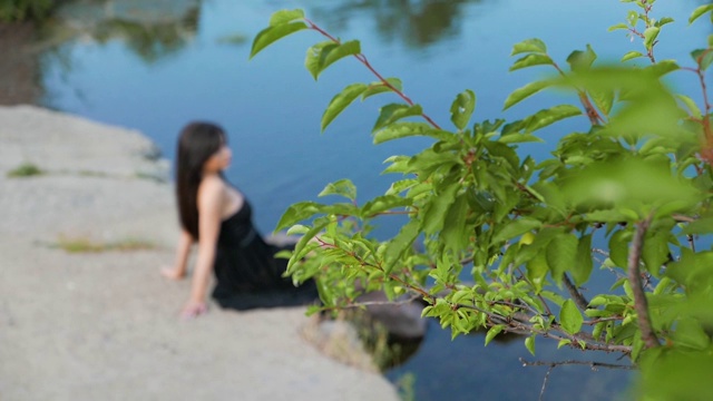 美丽的黑发长发的女孩，穿着黑色的连衣裙，坐在湖边的大石头上视频素材