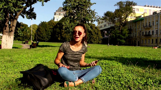 美丽的年轻女孩坐在公园的草地上，听着她最喜欢的电话。视频素材