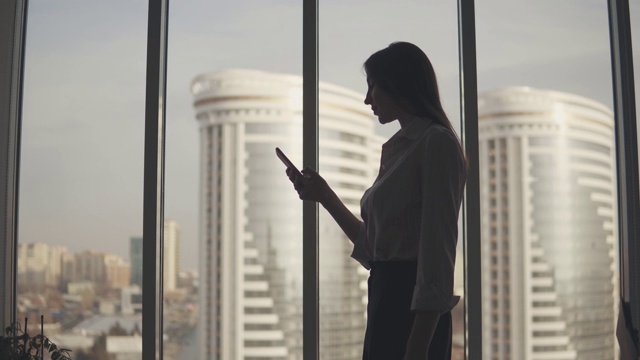 Silhouette of a young woman against the background of a large window and skyscraper. 女孩用智能手机。视频素材