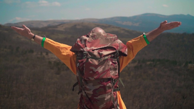 一个穿着黄色夹克、戴着眼镜、背着旅游背包的人站在群山中，高举着他的双手，这是自由的象征。视频素材