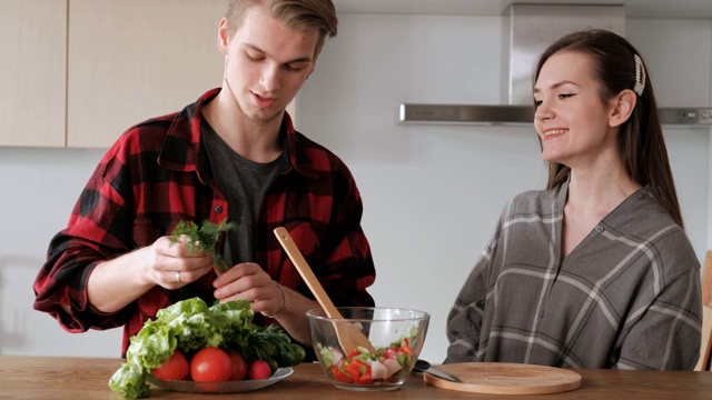 一对穿着格子衬衫的年轻漂亮夫妇正在厨房里做饭。一个女人和一个男人切蔬菜，在一个透明的玻璃盘子里用胡椒、番茄和萝卜做沙拉。视频素材