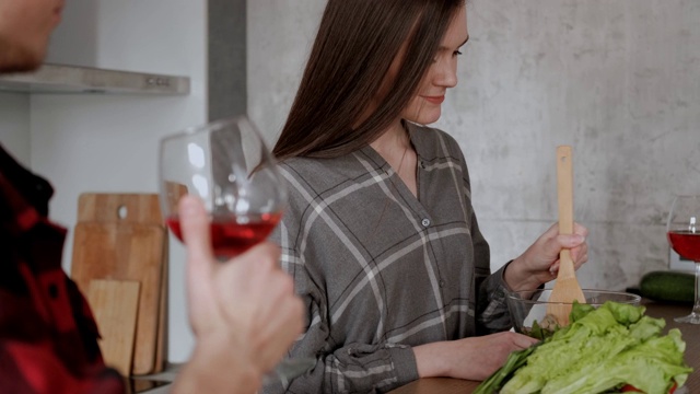 一对穿着格子衬衫的年轻漂亮夫妇正在厨房里做饭。一位女士和一位男士正在用大高脚杯喝红酒。视频素材