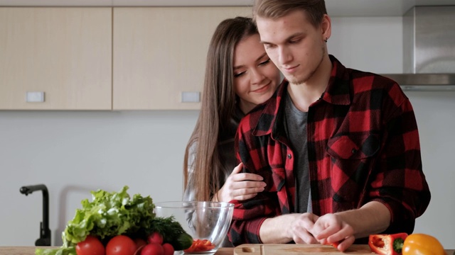 一对穿着格子衬衫的年轻漂亮夫妇正在厨房里做饭。一个女人和一个男人切蔬菜，在一个透明的玻璃盘子里用胡椒、番茄和萝卜做沙拉。视频素材