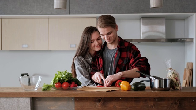 一对穿着格子衬衫的年轻漂亮夫妇正在厨房里做饭。一个女人和一个男人切蔬菜，在一个透明的玻璃盘子里用胡椒、番茄和萝卜做沙拉。视频素材