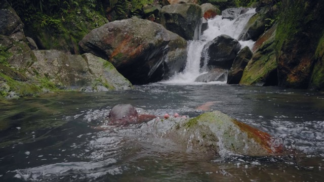 一名年轻人在热带森林瀑布流下的小溪中游泳。旅人在热带雨林的瀑布河中沐浴。视频素材