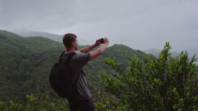 一个背着背包的旅行者站在山顶上用手机拍风景。视频下载