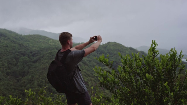 一个背着背包的旅行者站在山顶上用手机拍风景。视频下载