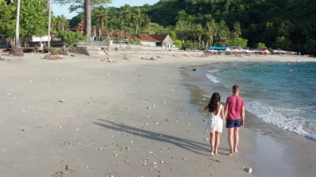 男人和女人沿着热带海滩在日落，跟踪航拍的年轻夫妇度假在海滩上玩。一对情侣漫步在空旷的海滩上，夕阳西下视频素材