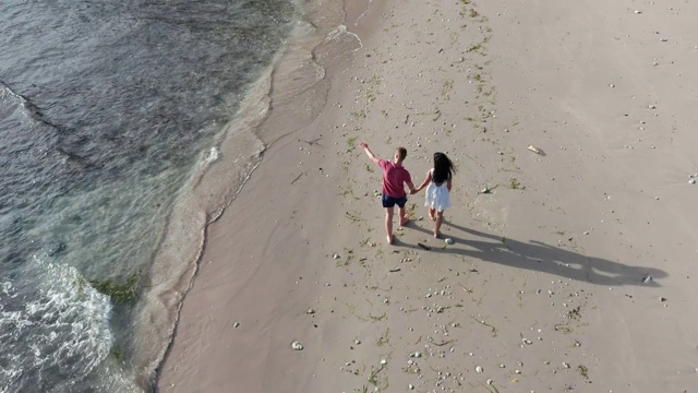 男人和女人沿着热带海滩在日落，跟踪航拍的年轻夫妇度假在海滩上玩。一对情侣漫步在空旷的海滩上，夕阳西下视频素材