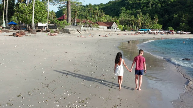 男人和女人沿着热带海滩在日落，跟踪航拍的年轻夫妇度假在海滩上玩。一对情侣漫步在空旷的海滩上，夕阳西下视频素材