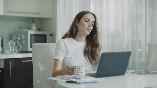 疲惫的女人在家里用笔记本电脑。在厨房喝茶的正经女人视频素材