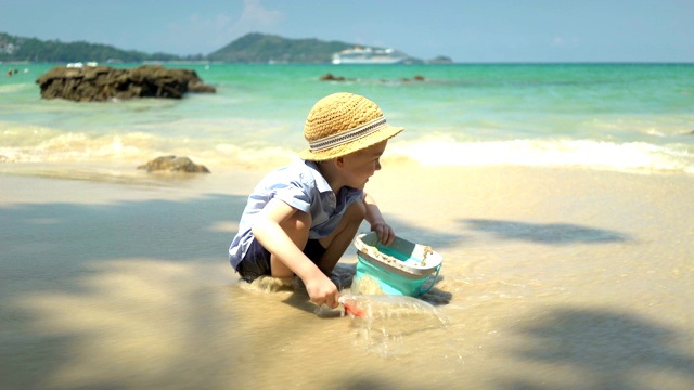 可爱的男孩在海边的沙滩上玩耍。他挖沙子，扔进桶里视频素材