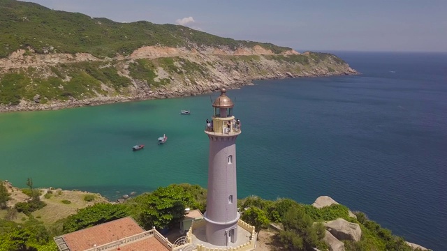 无人机看海灯塔在山崖和帆船。空中景观灯塔在绿色的山和船航行在绿松石海视频素材