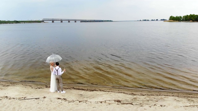 空中，美丽的新婚夫妇站在海滩上，拿着香槟，在透明的伞下，对着蓝天、河流和河上的大桥。春季的一天。婚礼视频素材