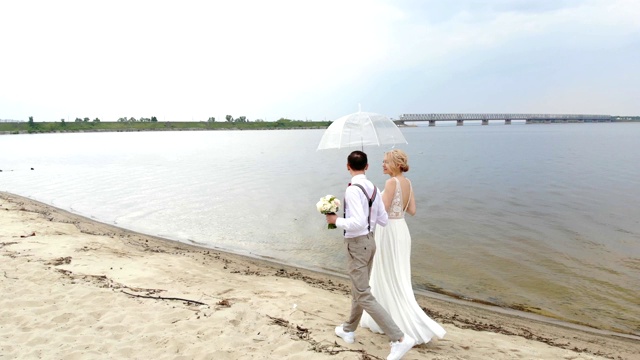 空中，美丽的新婚夫妇走在沙滩上，撑着一把透明的伞，对着蓝天、河流和河上的一座大桥。春天阳光明媚，有点下雨。婚礼视频素材