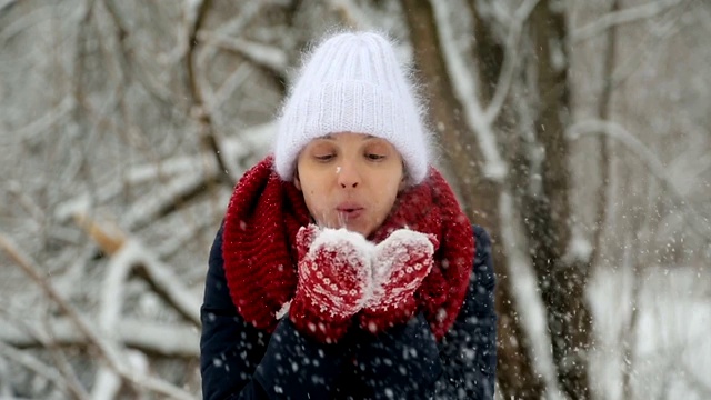 年轻的姑娘戴着白色的针织帽，戴着红色的围巾和手套吹雪视频素材