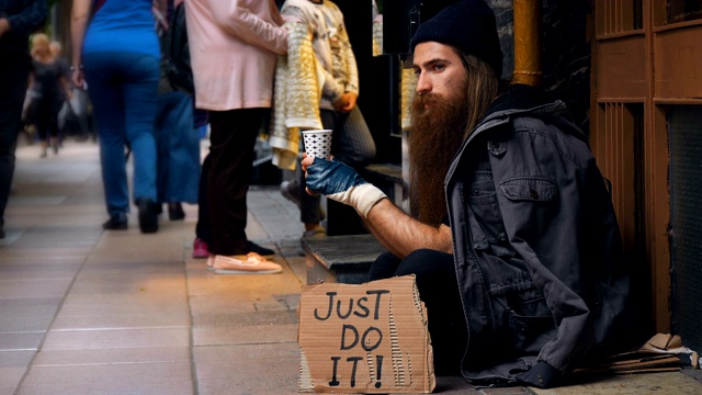 一个无家可归的人拿着“Just Do It”纸板，在拥挤的街道上乞讨视频素材