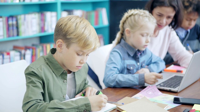 跟踪中景拍摄的中年教师和一群不同类型的小学生坐在教室的课桌前。导师向女学生解释如何在笔记本电脑上完成任务视频素材