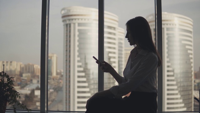 一个手持手机的女孩的剪影出现在现代建筑的背景上。商务女性在办公室使用智能手机。视频素材