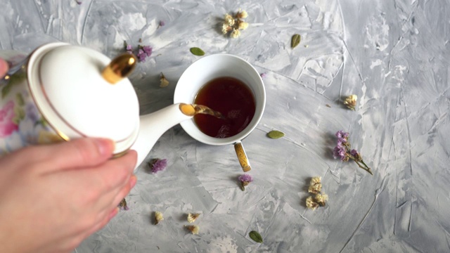 女人从白色茶壶里倒出棕色的茶，倒在白色的茶杯里，茶杯的背景是灰色的，有花视频下载