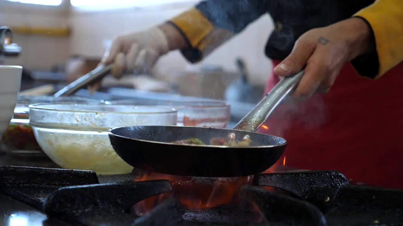 在酒店现代化的厨房里，不知名的厨师拿着两个平底锅，烹饪火焰风格的菜肴。男厨师在炉子上燃烧的煎锅里搅拌和煎炒蔬菜。烹饪的概念。慢莫侧视图视频素材