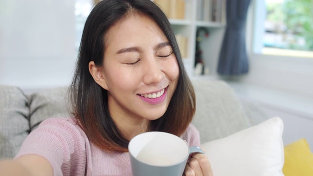 年轻的亚洲少女在家里做视频博客，女性喝着咖啡，在客厅用智能手机制作视频到社交媒体。生活方式女人在家里放松上午的概念。视频素材