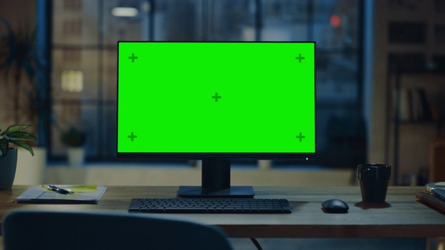 现代创意办公室的桌面电脑和绿色屏幕模型站在木桌上。在背景温暖的夜晚照明和开放空间工作室与城市窗户景观。缩小照片视频素材