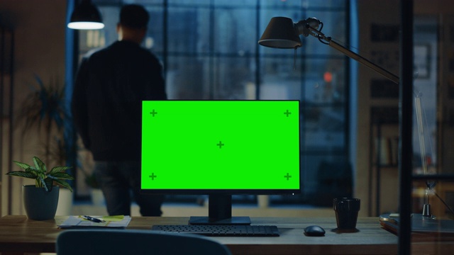 现代创意办公室的桌面电脑和绿色屏幕模型站在木桌上。在背景中，设计师从杯子里喝着饮料，看着窗外的夜城视频素材