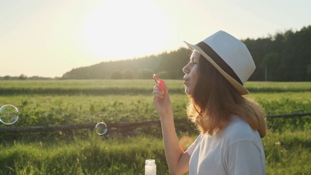 美丽的少女在一个帽子吹肥皂泡，夏天的自然景观背景视频素材