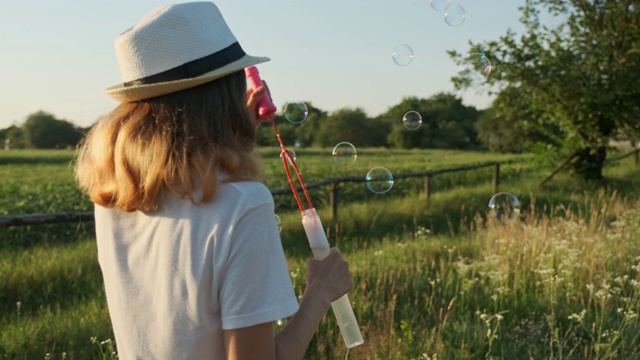 美丽的少女在一顶帽子吹肥皂泡，夏日的自然景观背景，黄金时刻视频素材