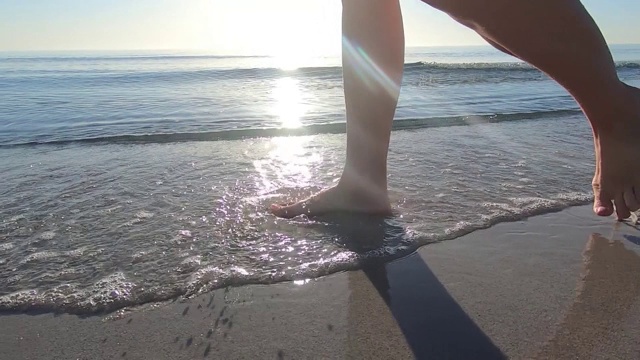 一个年轻女子的腿沿着海岸走着，海浪拍打着她的脚和沙滩，在黎明的背光下，前景是太阳视频素材