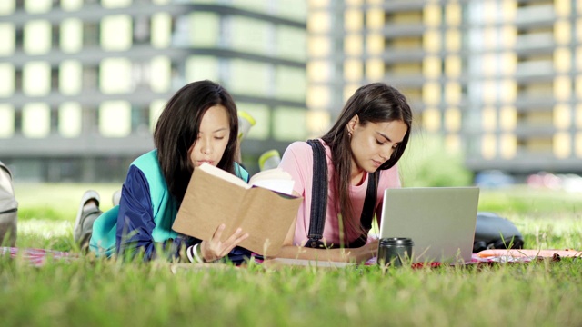 两个不同种族的女大学生躺在草地上的毯子上一起做项目，白人女孩在笔记本电脑上打字，她的亚洲朋友在读教科书视频素材