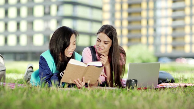 两个不同的女大学生在玩的时候躺在草地上的毯子上，一起做项目。快乐的亚洲女孩为她微笑的朋友在笔记本电脑上打字读书视频素材